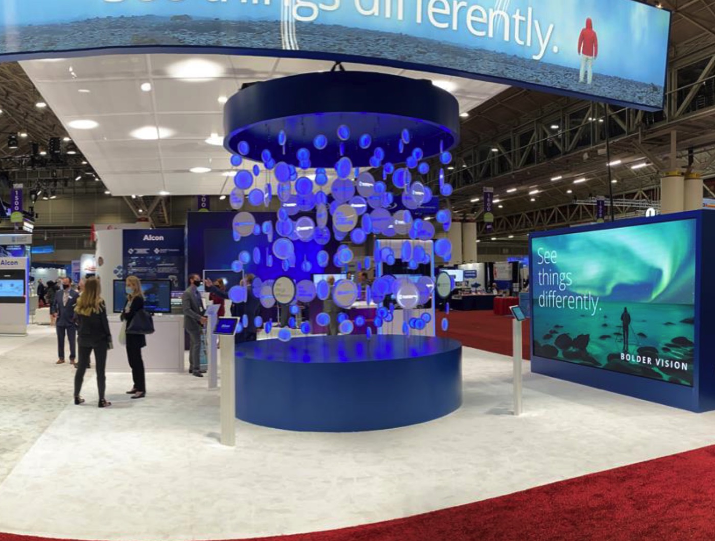 A two-story chandelier of glowing blue discs suspended over a circular booth stage on a busy trade show floor