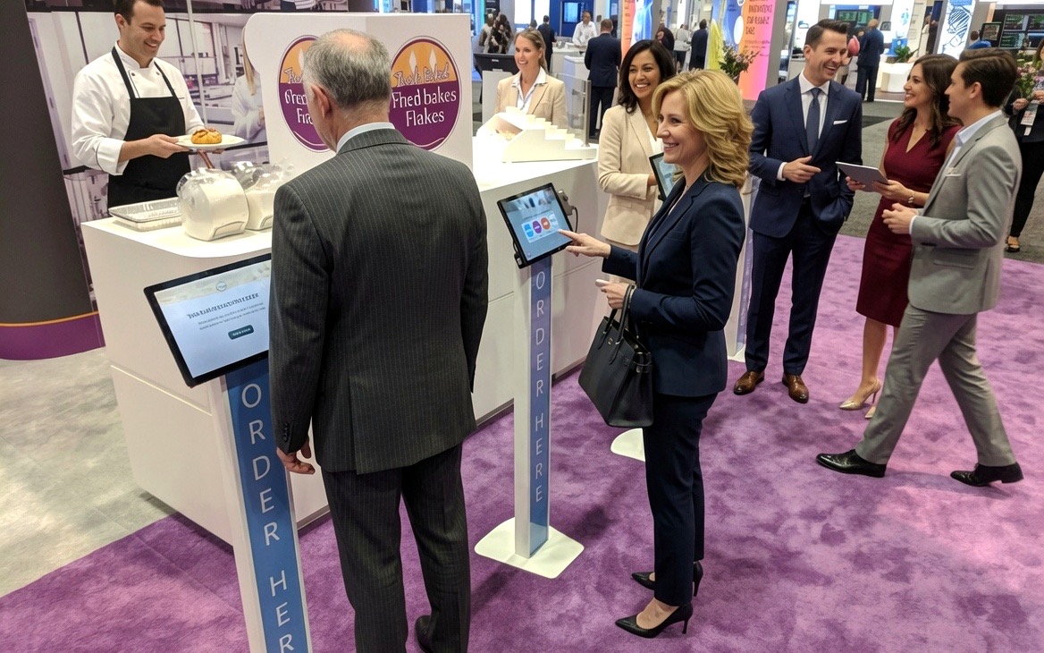 A chef serves fresh pastries beside self-serve ordering kiosks while attendees mingle on a purple show-floor carpet