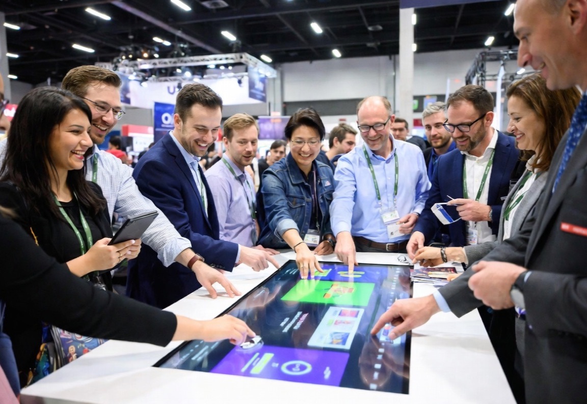 A crowd of laughing attendees plays a game together around a large interactive touch table on a trade show floor