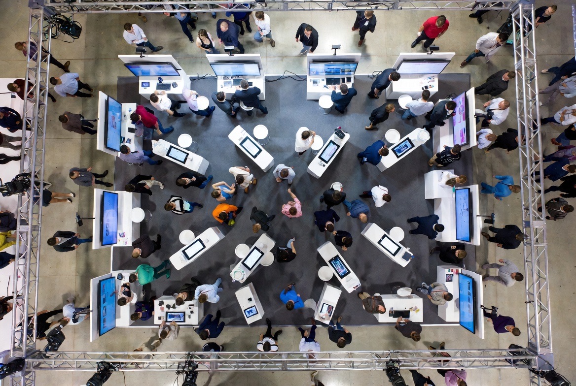 Aerial top-down view of a busy trade show booth: rings of white demo counters and screens inside a truss frame, with attendees filling the floor and the aisles on every side